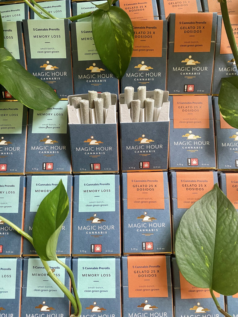 Magic Hour Prerolls packages lined up next to each other in blue and orange varieties in the center of the image the prerolled joints are positioned partially outside of the container and the display is adorned with leafy vines from the philodendron plant