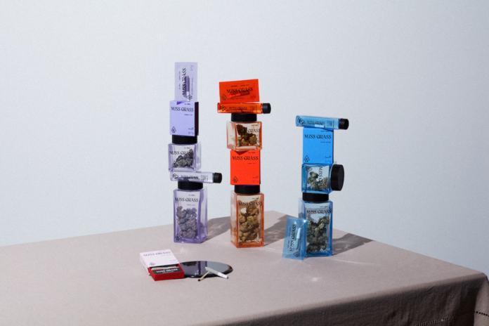 product display of Miss Grass flower a rolled join lays on a table in front of three stacks of weed filled jars, one purple, one red, and one blue