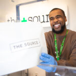The Source+ to Support Local Nonprofit, The Cupcake Girls, Throughout March budtender handing source + package to customer on the other side of the counter the budtender is wearing a brown long sleeve shirt and disposable gloves he is cheesin hard