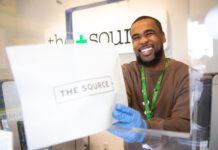 The Source+ to Support Local Nonprofit, The Cupcake Girls, Throughout March budtender handing source + package to customer on the other side of the counter the budtender is wearing a brown long sleeve shirt and disposable gloves he is cheesin hard