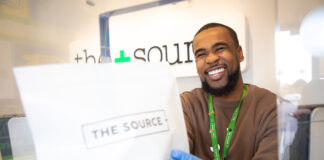 The Source+ to Support Local Nonprofit, The Cupcake Girls, Throughout March budtender handing source + package to customer on the other side of the counter the budtender is wearing a brown long sleeve shirt and disposable gloves he is cheesin hard