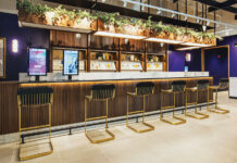 Cannabist Integrates Design and Technology in New Ways cannabist bar with navy blue walls hard wood floor velvet blue bar chairs with gold legs plants and lights hang down from the ceiling and touch screens line the bar