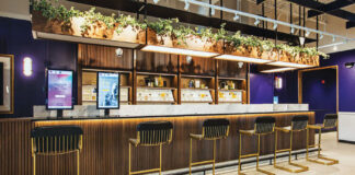 Cannabist Integrates Design and Technology in New Ways cannabist bar with navy blue walls hard wood floor velvet blue bar chairs with gold legs plants and lights hang down from the ceiling and touch screens line the bar