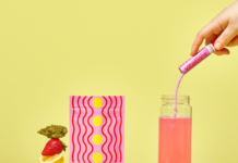 Cannabis-Infused Powdered Energy Drink Mix Launches: Vibations High + Energy neon yellow background display of pink drink mix container and weed nugs and a bottle of water with a white hand pouring pink drink mix into bottle