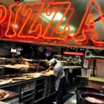 NY Considers Cannabis-Infused Food Sales at Pizzerias and Other Eateries Red neon pizza sign above a man making pizzas at a shop