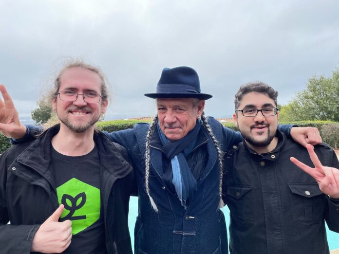 photo of three men smiling with their arms around one another. The middle man has two long gray braids and is wearing a fedora the man on the left is giving a thumbs up and the man on the right is throwing up a peace sign