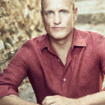 Woody Harrelson is Emerald Cup’s Willie Nelson Award Honoree for 2022 photo of Woody Harrelson with a bald head and a serious expression wearing a muted red shirt he is sitting on outdoor steps and leaning toward the camera with his elbows resting on his knees