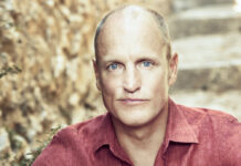 Woody Harrelson is Emerald Cup’s Willie Nelson Award Honoree for 2022 photo of Woody Harrelson with a bald head and a serious expression wearing a muted red shirt he is sitting on outdoor steps and leaning toward the camera with his elbows resting on his knees