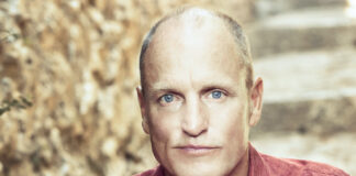 Woody Harrelson is Emerald Cup’s Willie Nelson Award Honoree for 2022 photo of Woody Harrelson with a bald head and a serious expression wearing a muted red shirt he is sitting on outdoor steps and leaning toward the camera with his elbows resting on his knees