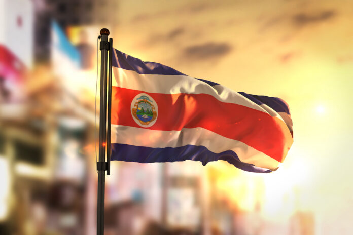Costa Rica Flag Against City Blurred Background At Sunrise Backlight 3D Rendering