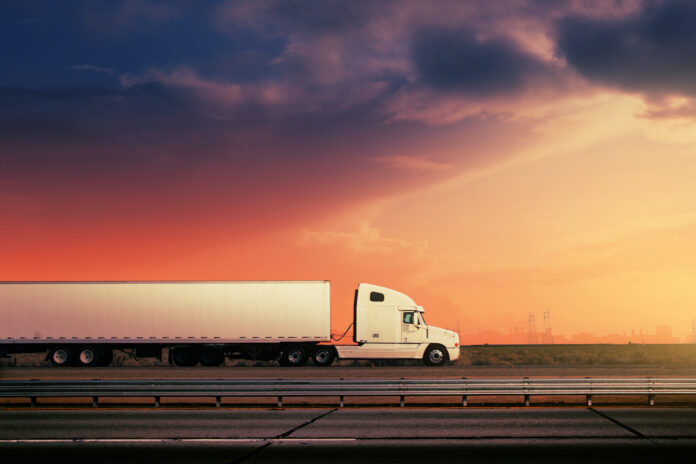 White freight truck driving on freeway road under red sunset sky.