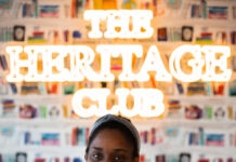 The Heritage Club Becomes First Black Woman Owned Cannabis Dispensary in Boston photo of founder nike john inside the heritage club dispensary she is sitting down and smiling and a heritage club neon sign shines behind her