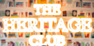 The Heritage Club Becomes First Black Woman Owned Cannabis Dispensary in Boston photo of founder nike john inside the heritage club dispensary she is sitting down and smiling and a heritage club neon sign shines behind her