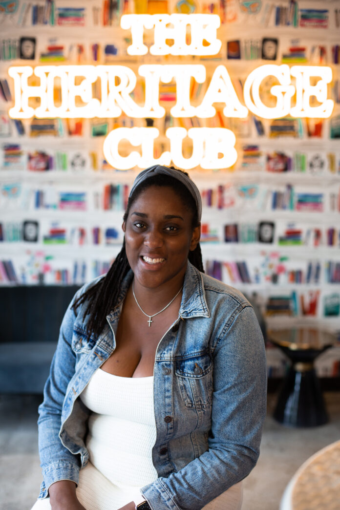 photo of founder nike john inside the heritage club dispensary she is sitting down and smiling and a heritage club neon sign shines behind her