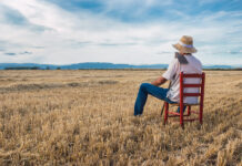 No-Till Farming Pays Dividends Farmer with straw hat, white t-shirt, and blue jeans sitting on a red chair surveying his land with a till on his shoulder