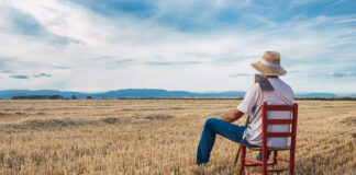 No-Till Farming Pays Dividends Farmer with straw hat, white t-shirt, and blue jeans sitting on a red chair surveying his land with a till on his shoulder
