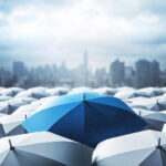 Identifying Key Tools to Survive an Economic Downturn Blue umbrella on top of other gray umbrellas on city background. Business and safety concept