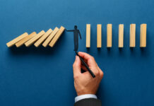 An Ounce of Prevention Can Stop Financial Distress Wide view image of male hand drawing silhouette of a man making a stop gesture to prevent wooden dominos from collapsing. Over navy blue background.