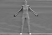 Cannabis Trademarks May Require a Tobacco Disguise 3D rendering of a woman trying to blend in with the black and white striped background, afraid to show her true colours. She is standing with her back against the wall and hiding like a wallflower.
