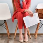 86% of Employers Plan to Hire More in 2024, but Benefits, DEI Declining young woman with modern red suit sitting for a job interview white chairs brown leather business bag