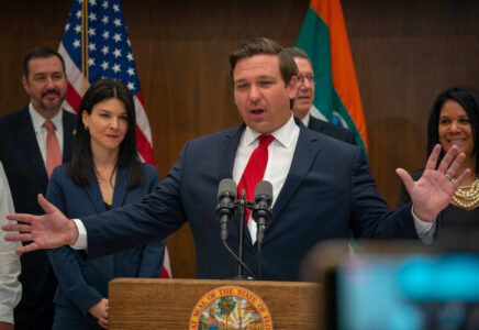 Florida Governor Ron DeSantis speaking in front of microphones behind a podium wearing a dark suit, white shirt, and red tie