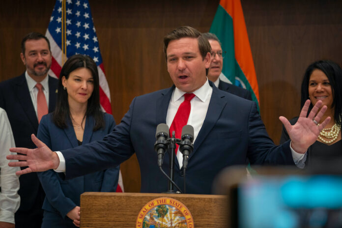 Florida Governor Ron DeSantis speaking in front of microphones behind a podium wearing a dark suit, white shirt, and red tie