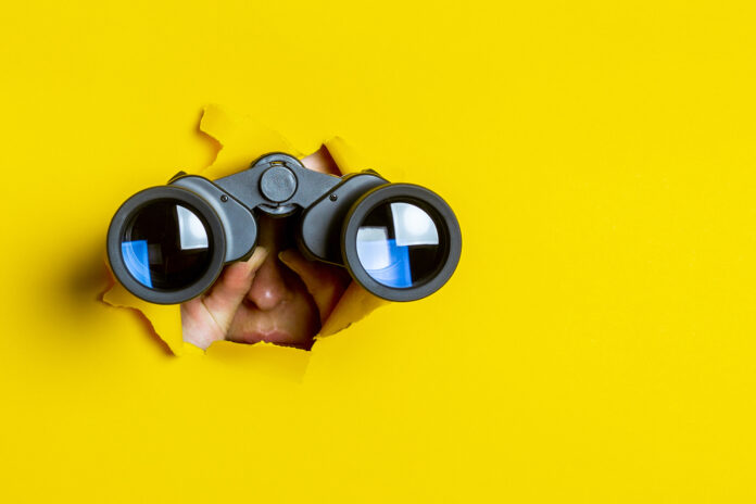 Female popping through a yellow banner with binoculars to spy as a secret shopper