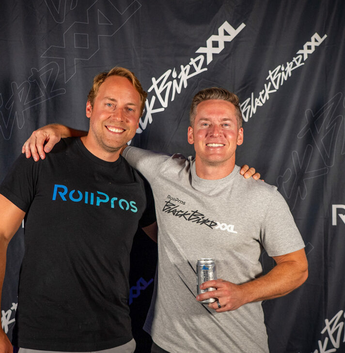 Two men pose in front of RollPros Blackbird XXL step-and-repeat banner