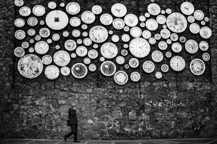 Black-and-white photo of a man walking past a stone wall covered in dozens of clocks, symbolizing the long history of cannabis prohibition.