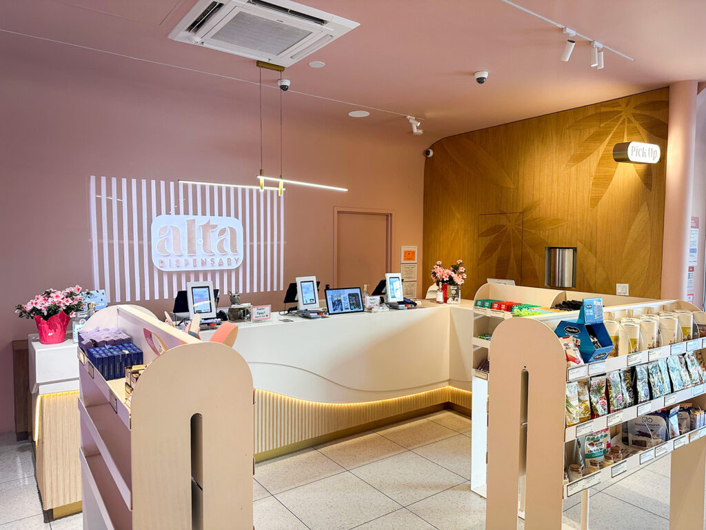 Checkout counter and retail product displays inside Alta Dispensary, featuring the Alta logo projection and wall art.