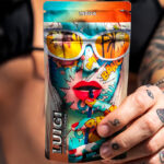 Premium Cannabis Meets Science and Style in Luigi Oil’s Flavor-Driven Lineup A tattooed woman in a black bikini holding a colorful Luigi Oil vape pouch featuring comic-style artwork and the word "Indica."