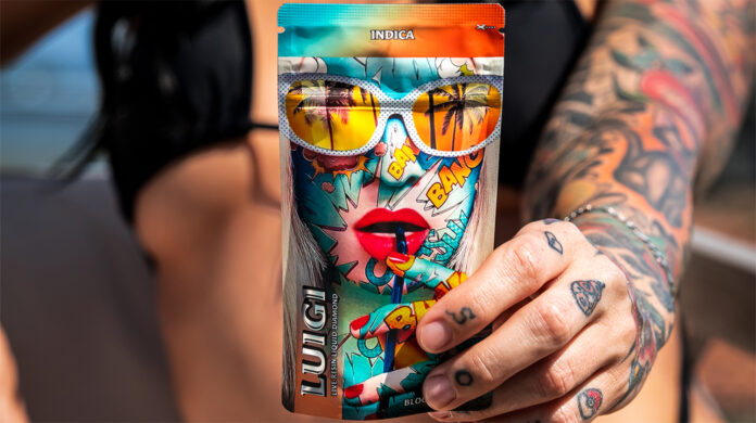 A tattooed woman in a black bikini holding a colorful Luigi Oil vape pouch featuring comic-style artwork and the word "Indica."