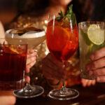 Alcohol Sales Slide While Cannabis Drinks Surge Toward $2.8B Market Four people clink colorful sparkling beverages in elegant glassware at a warm, upscale setting, symbolizing the shift toward low- and no-alcohol and cannabis-infused drink trends.
