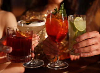 Alcohol Sales Slide While Cannabis Drinks Surge Toward $2.8B Market Four people clink colorful sparkling beverages in elegant glassware at a warm, upscale setting, symbolizing the shift toward low- and no-alcohol and cannabis-infused drink trends.