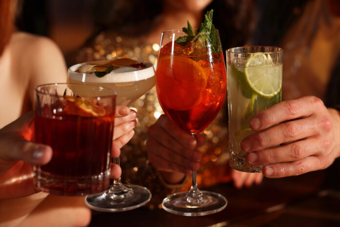 cannabis-drinks-rise-declining-alcohol-sales-toasting-glasses Four people clink colorful sparkling beverages in elegant glassware at a warm, upscale setting, symbolizing the shift toward low- and no-alcohol and cannabis-infused drink trends.