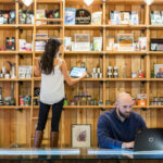 Dispensary Loss Prevention: How SOPs Stop Shrinkage A dispensary employee checks product inventory on a tablet while another works at a laptop behind the retail counter, illustrating back-of-house SOPs and operational oversight.