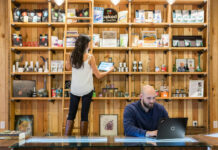 Dispensary Loss Prevention: How SOPs Stop Shrinkage A dispensary employee checks product inventory on a tablet while another works at a laptop behind the retail counter, illustrating back-of-house SOPs and operational oversight.