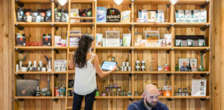 Dispensary Loss Prevention: How SOPs Stop Shrinkage A dispensary employee checks product inventory on a tablet while another works at a laptop behind the retail counter, illustrating back-of-house SOPs and operational oversight.