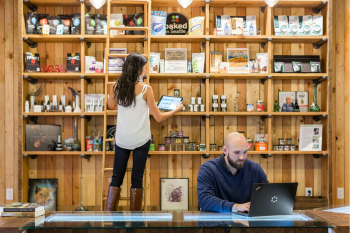 A dispensary employee checks product inventory on a tablet while another works at a laptop behind the retail counter, illustrating back-of-house SOPs and operational oversight.