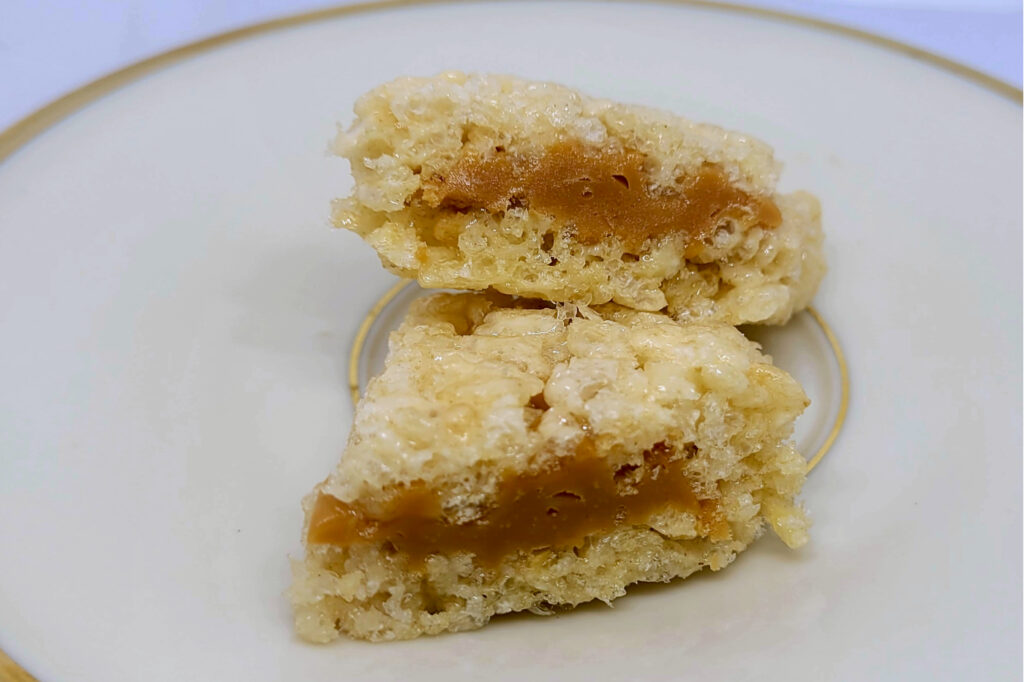 Close-up of a caramel-filled crispy rice square broken in half on a plate, showing gooey caramel and toasted cereal texture.
