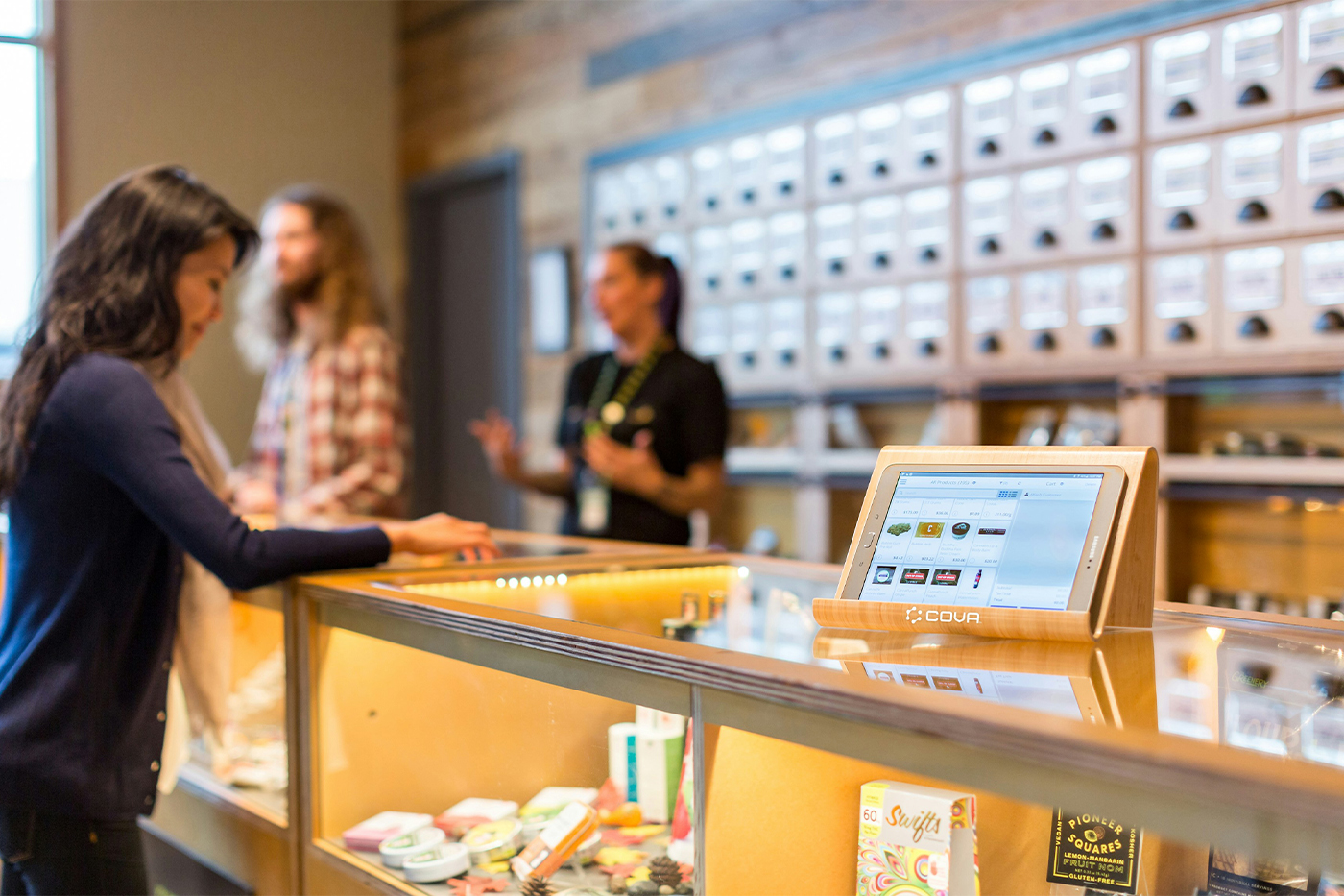 How Curated Menus Drive Cannabis Retail Differentiation Customer browsing products at a modern cannabis dispensary counter with a digital menu display in the foreground and staff assisting shoppers in the background.