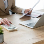 Business professional reviewing documents and using a calculator and laptop for financial reconciliation