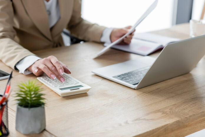 Business professional reviewing documents and using a calculator and laptop for financial reconciliation