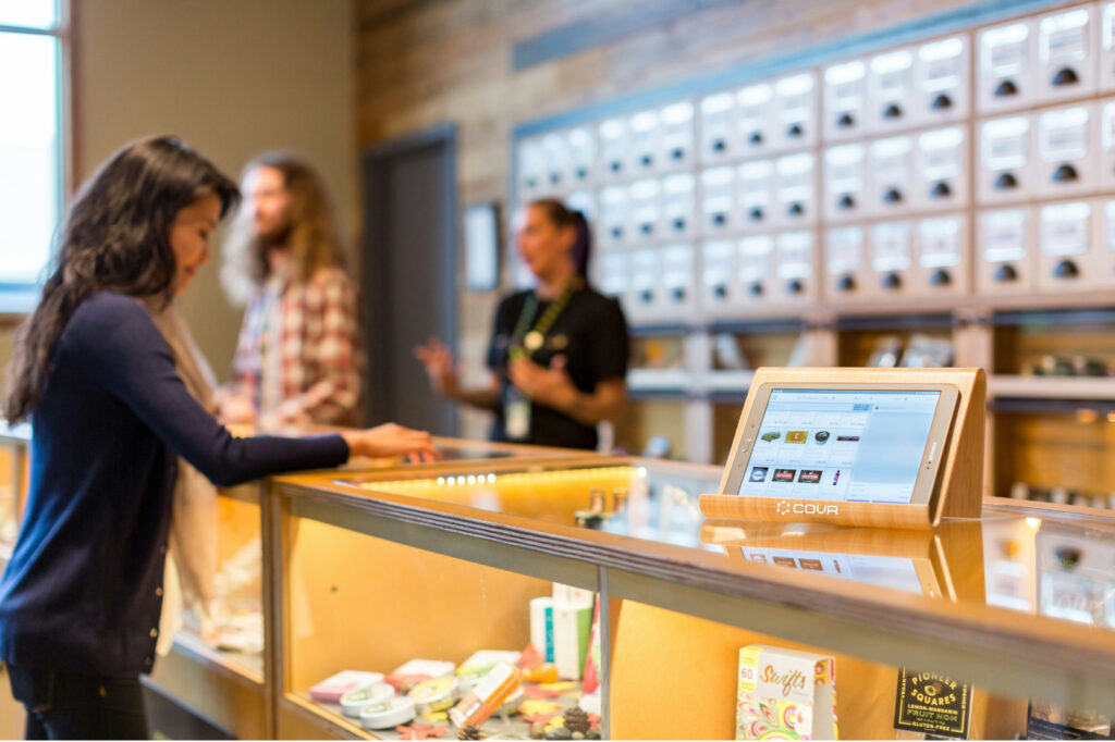 A customer stands at a dispensary counter with a point-of-sale tablet in the foreground.