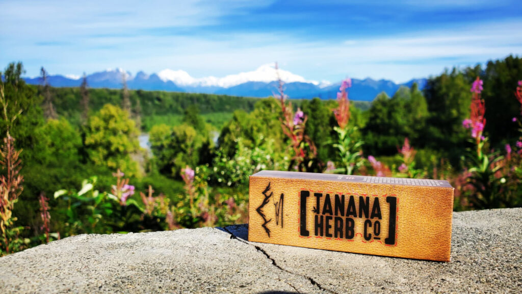 A Tanana Herb Co. product package sits outdoors with an Alaska landscape in the background.