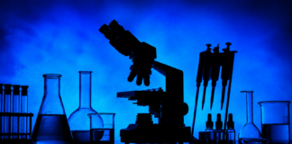 Rescheduling Cannabis: The Coming Collision with Ag-Biotech Regulation Silhouetted laboratory microscope, glass beakers, test tubes, pipettes, and sample bottles against a blue background for a cannabis biotechnology research story.