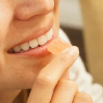 Edibles Are Growing Up, and Manufacturing Needs to Catch Up Close-up profile of a smiling woman holding a single square peach-colored cannabis gummy candy to her lips.