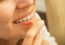 Edibles Are Growing Up, and Manufacturing Needs to Catch Up Close-up profile of a smiling woman holding a single square peach-colored cannabis gummy candy to her lips.