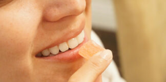 Edibles Are Growing Up, and Manufacturing Needs to Catch Up Close-up profile of a smiling woman holding a single square peach-colored cannabis gummy candy to her lips.
