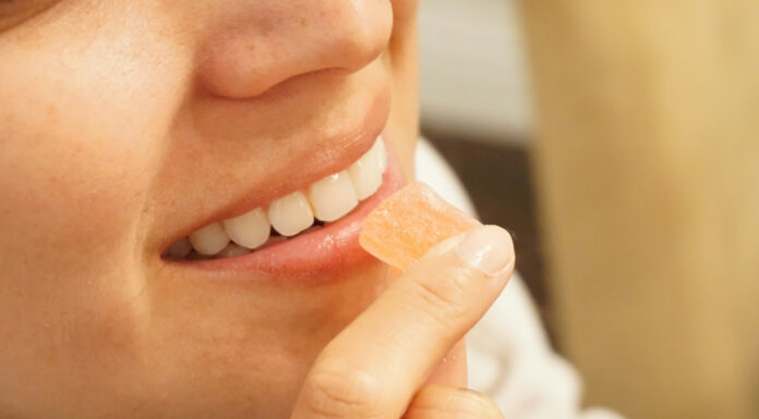 Edibles Are Growing Up, and Manufacturing Needs to Catch Up Close-up profile of a smiling woman holding a single square peach-colored cannabis gummy candy to her lips.